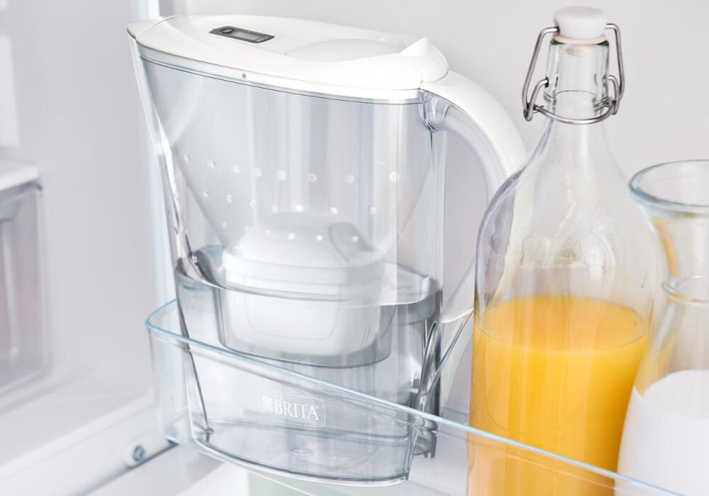 Dzbanek filtrujący z przezroczystego plastiku z białą pokrywką znajduje się na półce w lodówce obok butelki z sokiem pomarańczowym. Na dzbanku widoczne jest logo \'BRITA\'.