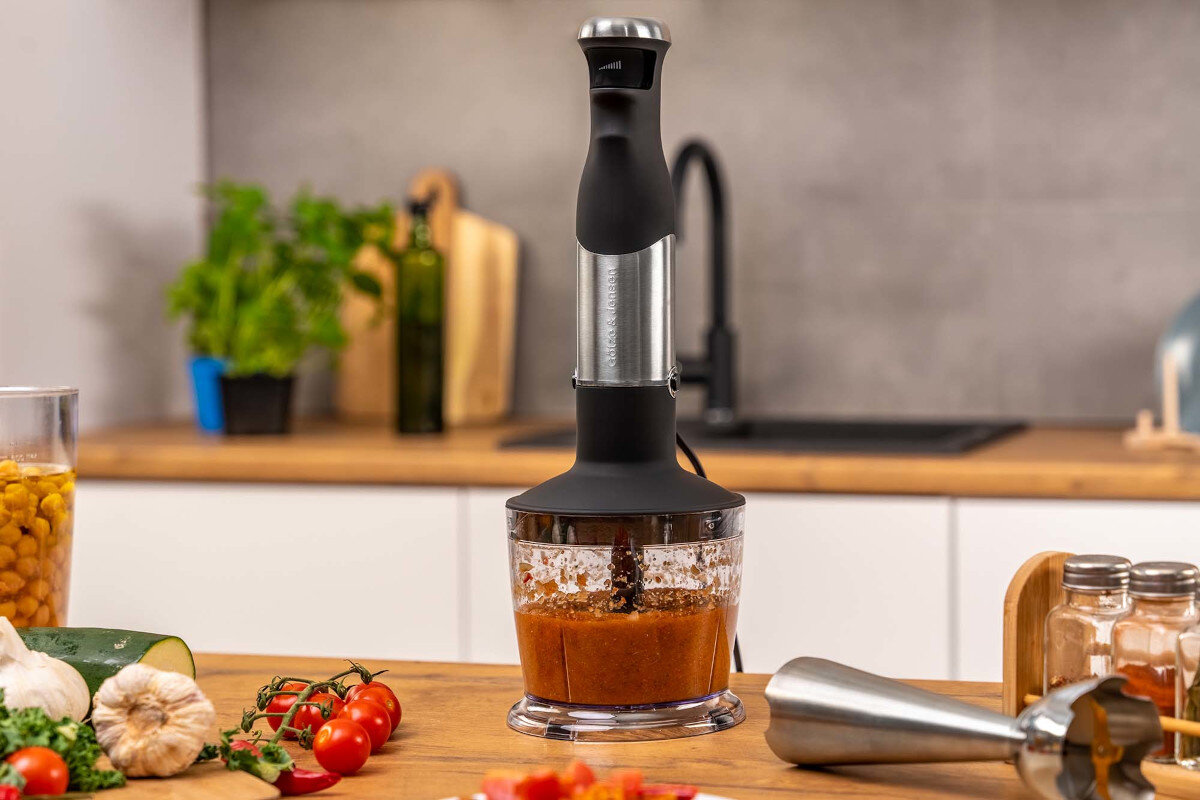 Na blacie kuchennym stoi blender z czarną rączką i przezroczystym pojemnikiem, w którym znajduje się zmiksowana pomarańczowa substancja. W tle widoczne są zioła w doniczce, oliwa oraz inne akcesoria kuchenne.