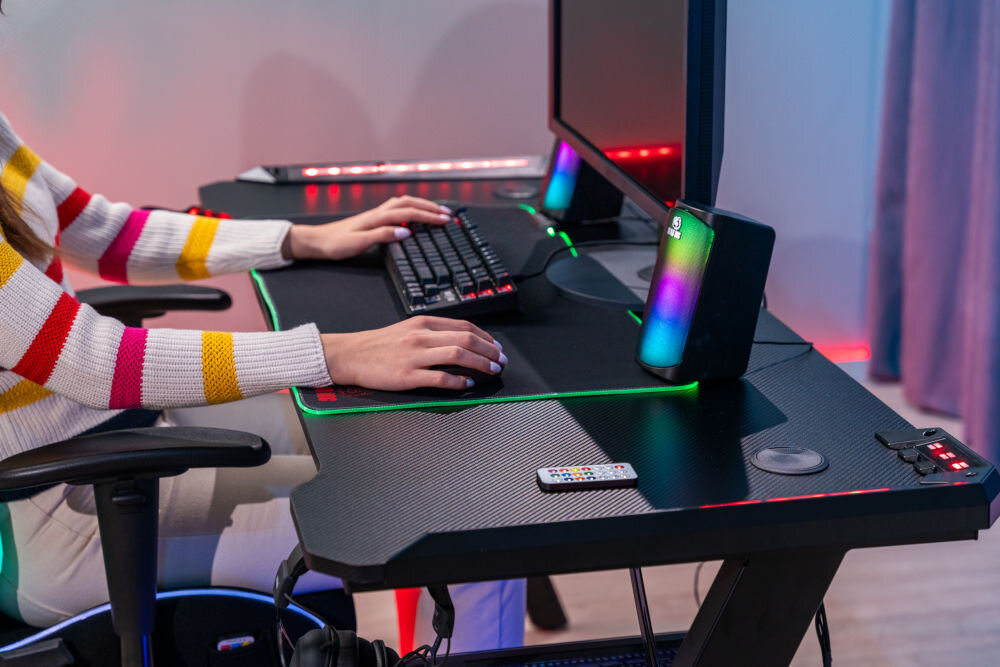 Biurko gamingowe z klawiaturą, myszką i monitorem. Na biurku znajduje się pilot oraz głośniki z podświetleniem RGB.