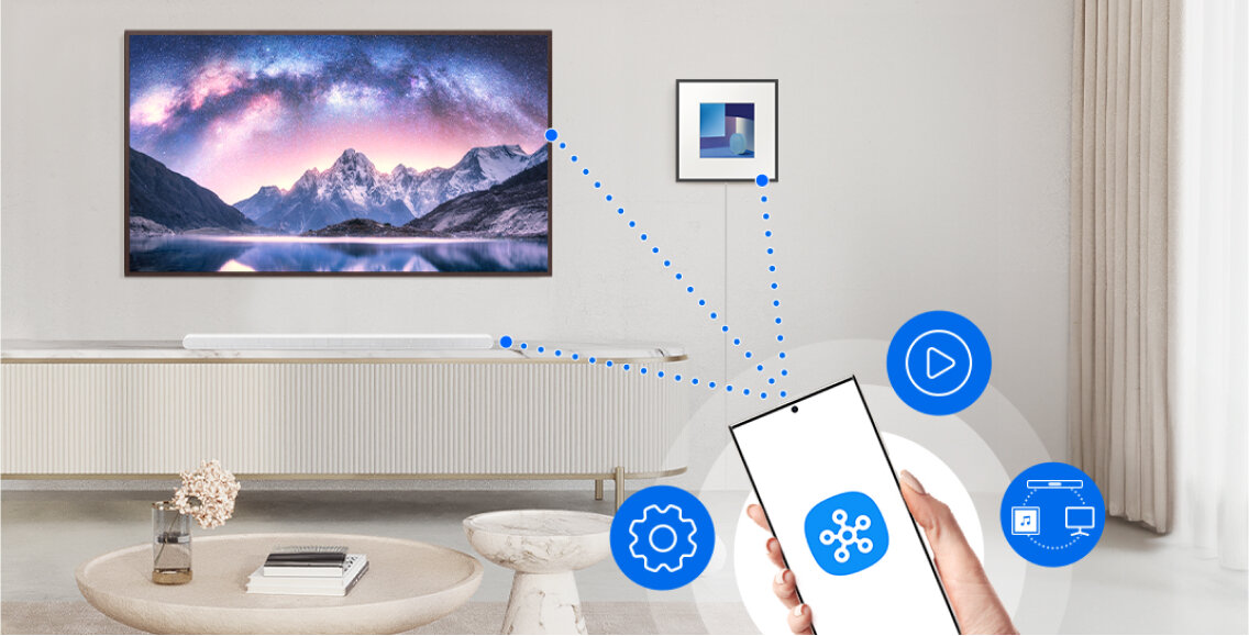 W salonie znajduje się duży telewizor z krajobrazem górskim na ekranie oraz biały soundbar pod nim. Na pierwszym planie widać rękę trzymającą smartfon z ikoną aplikacji, a wokół niej są ikony ustawień, odtwarzania i połączenia z urządzeniami.