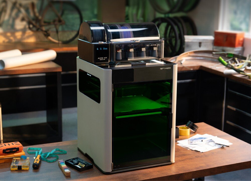 Drukarka 3D BAMBU LAB H2D Laser Full Combo Drukarka 3D z górnym podajnikiem stojąca na blacie w warsztacie z narzędziami