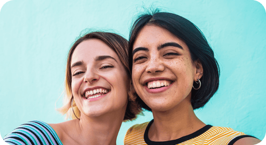 Dwie uśmiechnięte kobiety robią selfie smartfonem. Dwie osoby uśmiechają się na tle jasnoniebieskiej ściany. Obie mają krótkie włosy i noszą kolorowe ubrania.