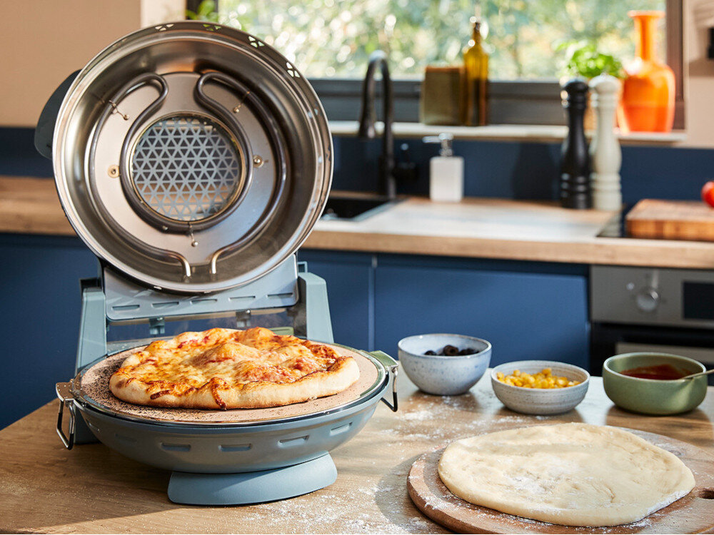 Na blacie kuchennym znajduje się otwarty piec do pizzy z gotową pizzą w środku. Obok leży surowe ciasto na desce oraz miseczki z dodatkami.