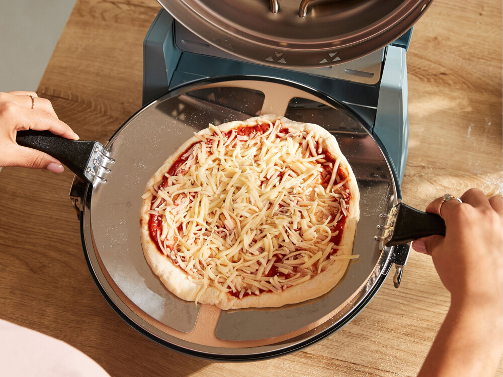 Osoba trzyma łopatkę z surową pizzą pokrytą serem i sosem pomidorowym, przygotowaną do włożenia do pieca do pizzy. Piec ma metaliczną obudowę i znajduje się na drewnianym blacie.