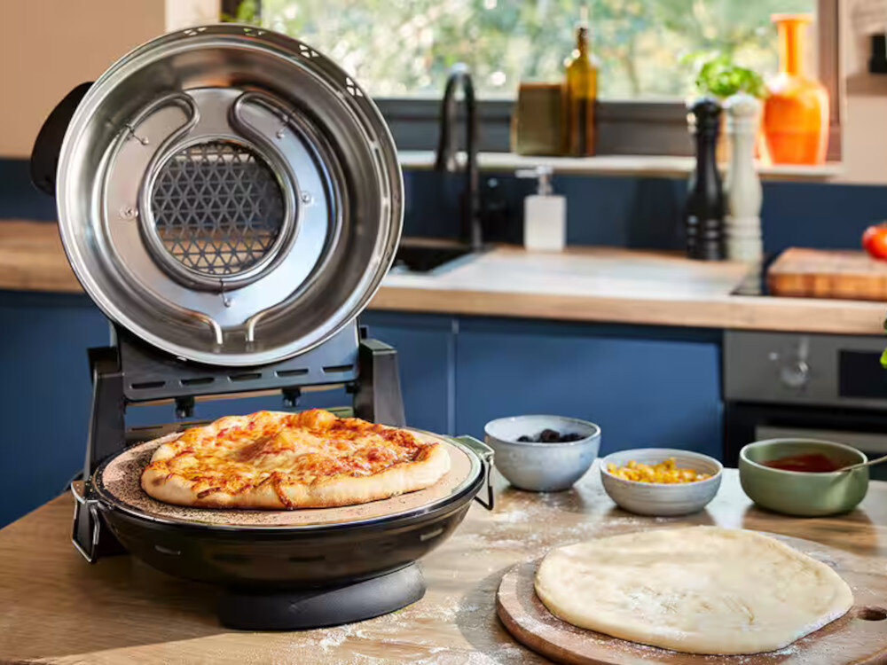 Na blacie kuchennym znajduje się otwarty piec do pizzy z gotową pizzą w środku. Obok leży surowe ciasto na desce oraz miseczki z dodatkami.