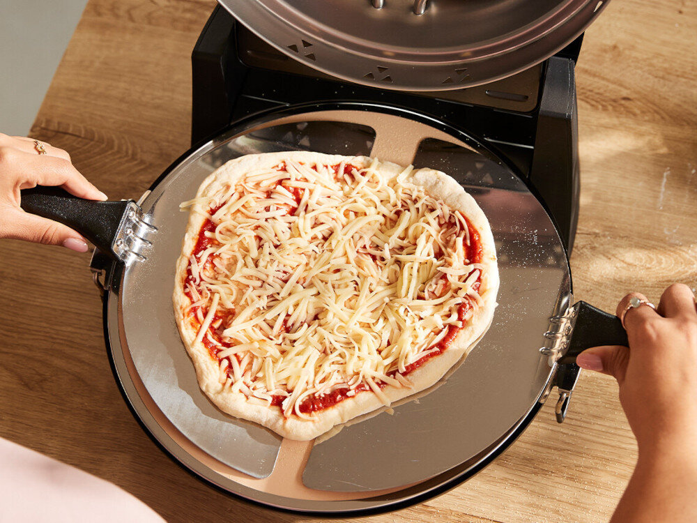 Osoba trzyma łopatkę z surową pizzą pokrytą sosem pomidorowym i startym serem, umieszczoną na metalowej powierzchni pieca do pizzy. Piec znajduje się na drewnianym blacie.