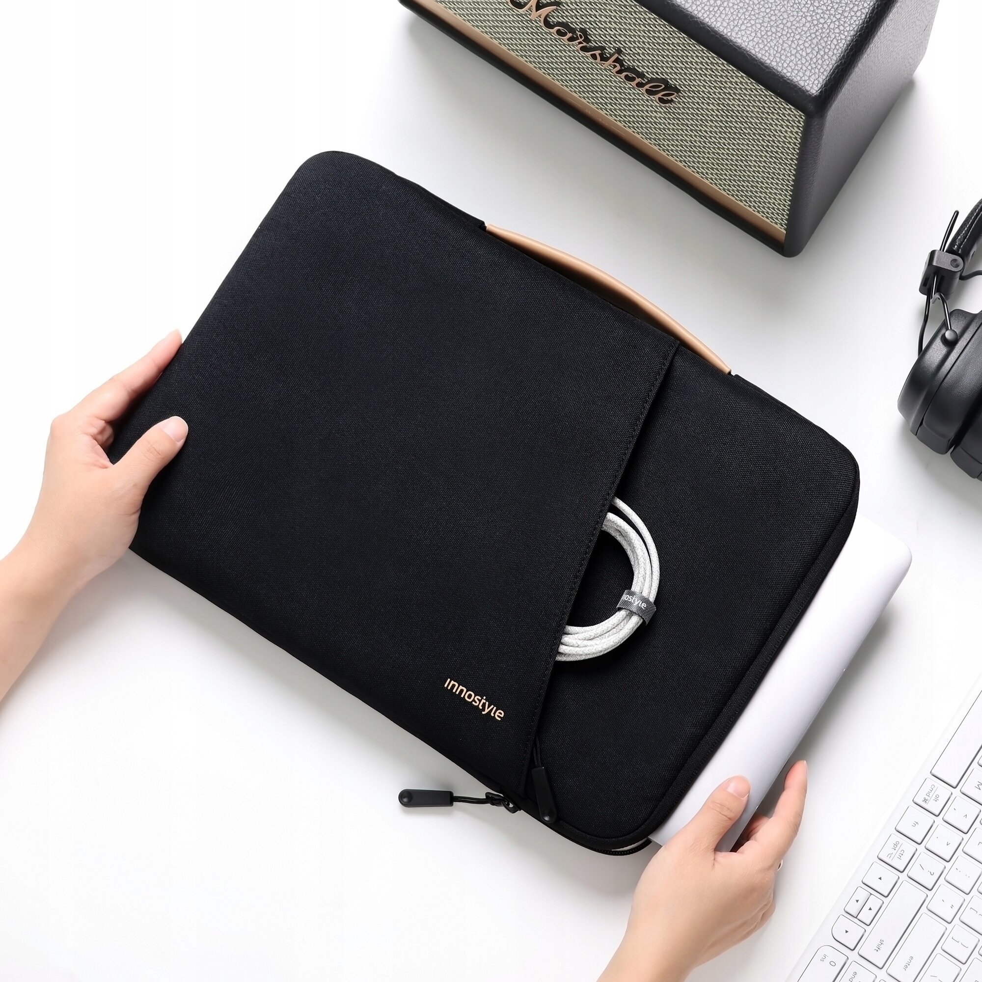 Czarna torba na laptopa z napisem \'innostyle\', trzymana przez dwie ręce, z widocznym białym kablem i laptopem częściowo wsuniętym do środka.