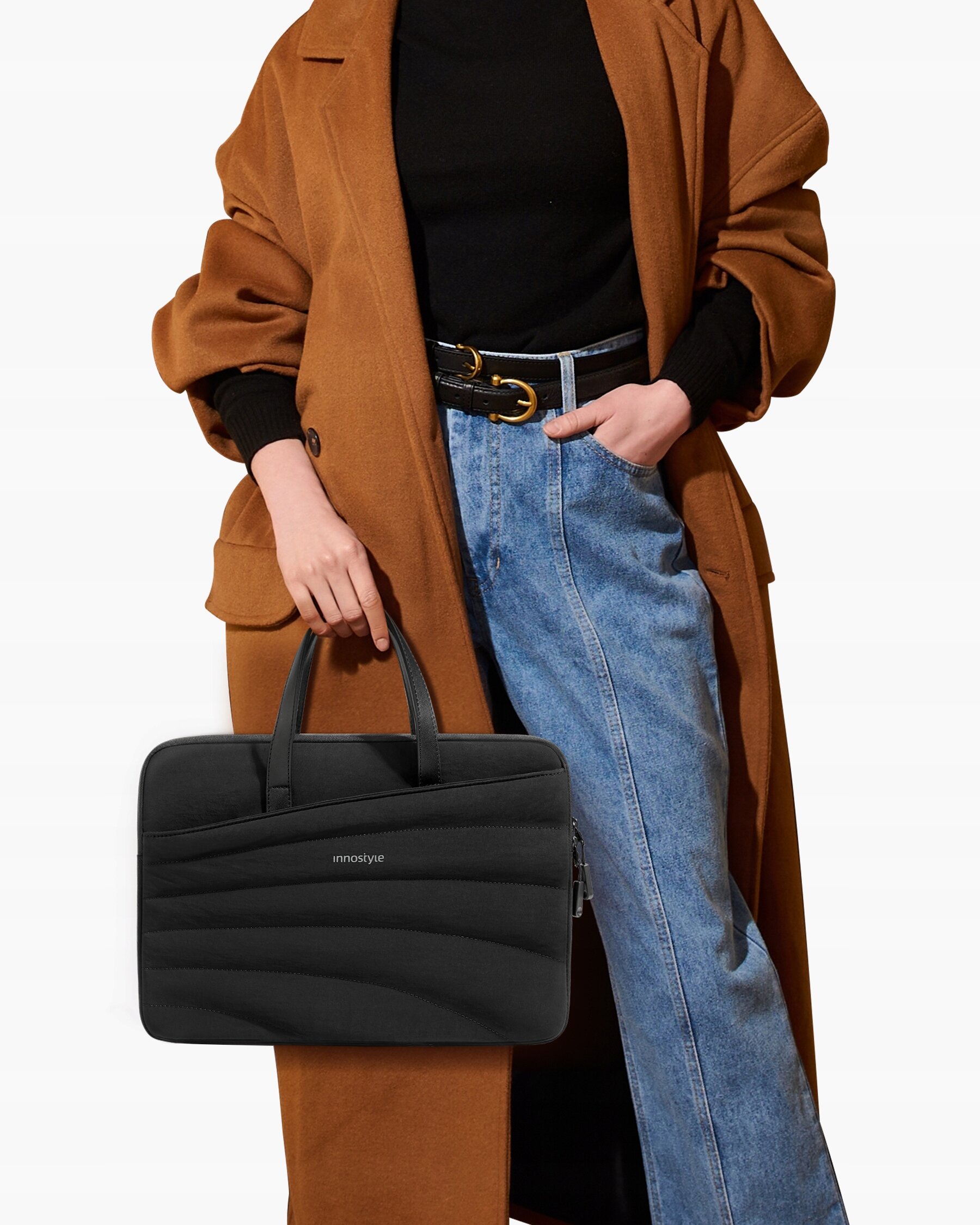 Czarna torba na laptopa marki Innostyle trzymana przez osobę ubraną w brązowy płaszcz i niebieskie jeansy. Na torbie widoczny jest napis \'innostyle\'.