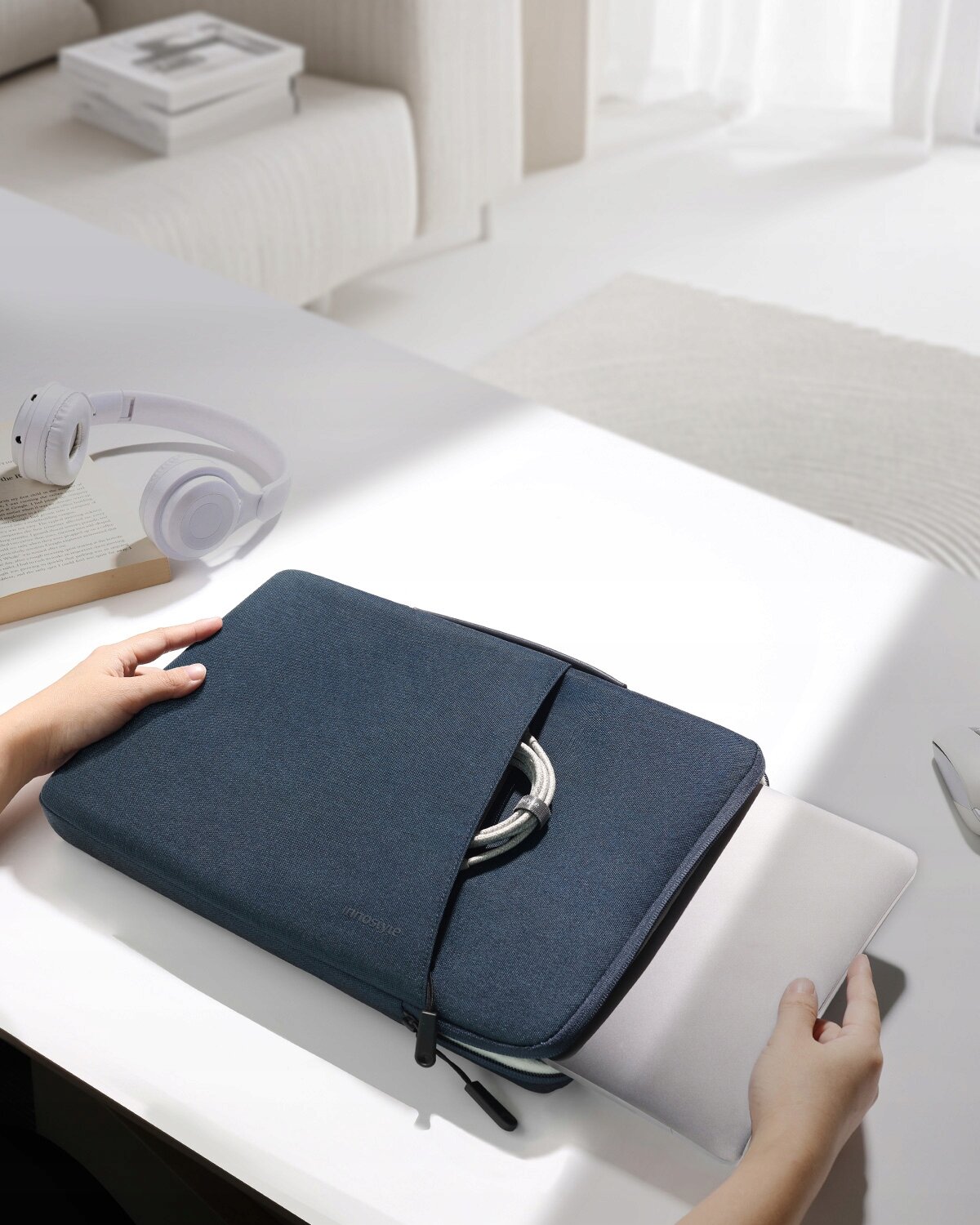 Niebieska torba na laptopa z suwakiem, w której znajduje się laptop, leży na białym stole obok białych słuchawek i książki. Na torbie widoczny jest napis \'Innostyle\'.
