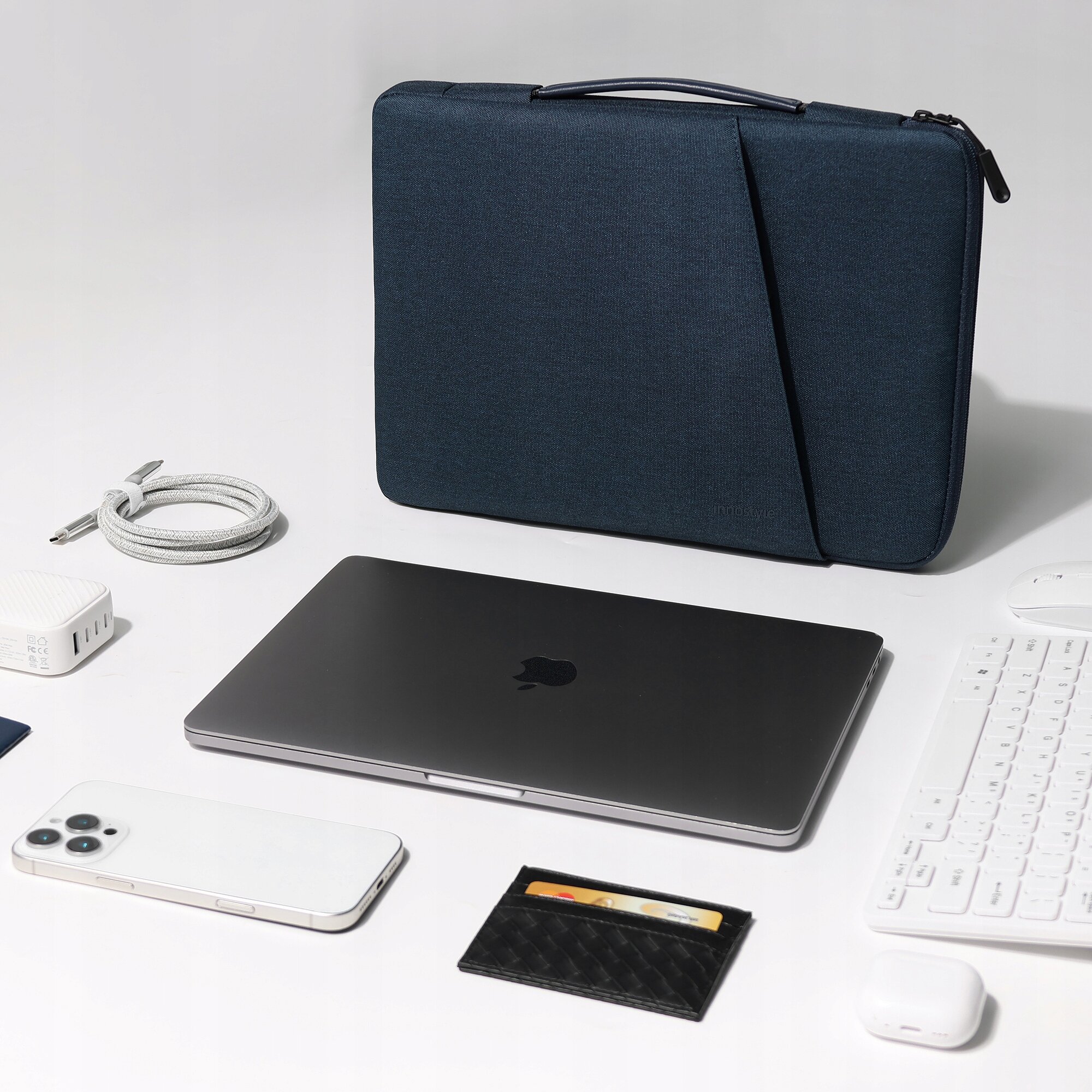 Na zdjęciu znajduje się niebieska torba na laptopa stojąca pionowo na białym tle, obok której leży laptop, smartfon, klawiatura, kabel, ładowarka, portfel i słuchawki. Torba ma prosty, elegancki design z zamkiem błyskawicznym na górze.