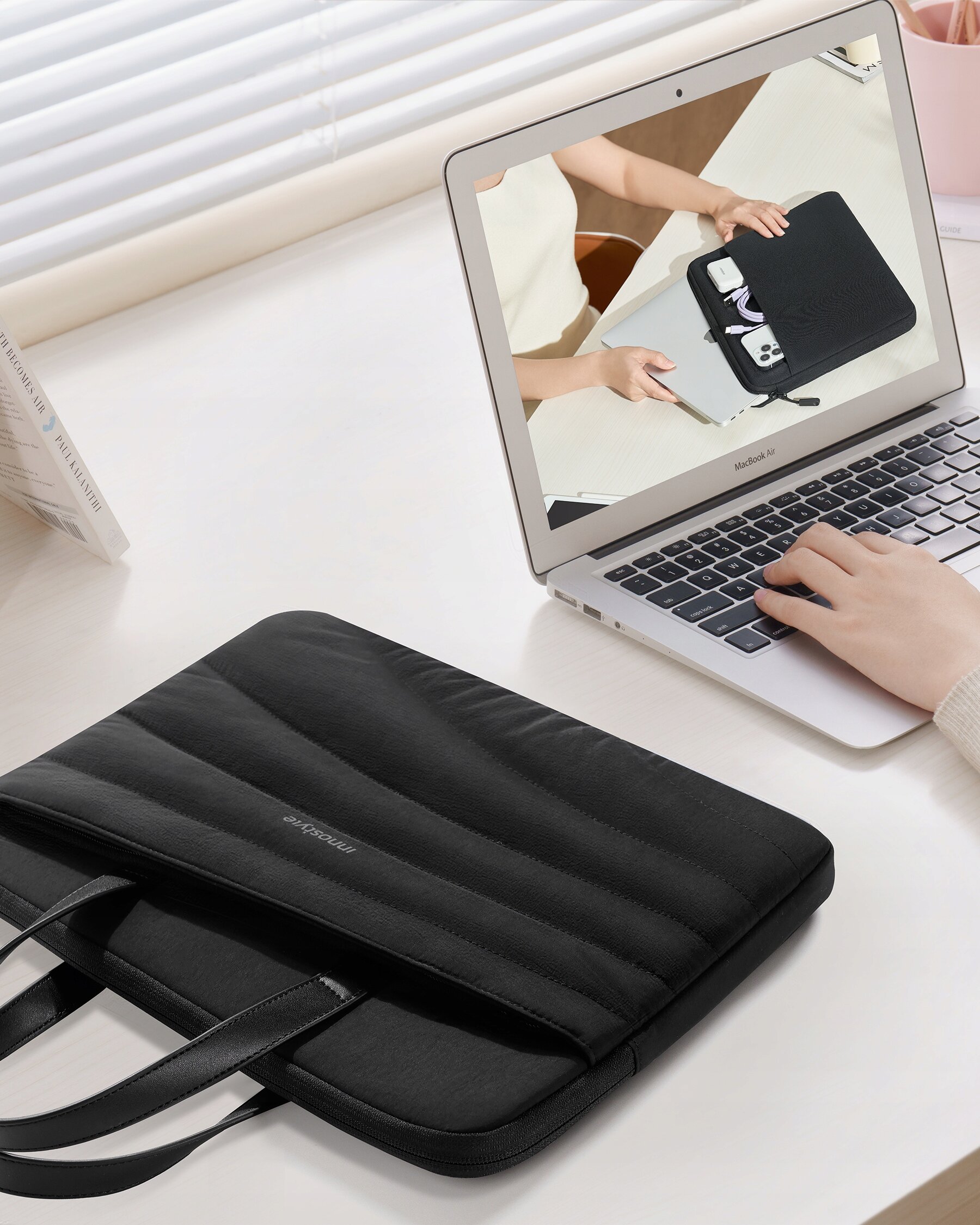 Czarna torba na laptopa leży na biurku obok otwartego laptopa, na którego ekranie widoczna jest osoba wkładająca przedmioty do podobnej torby. Na torbie widoczny jest napis \'INNOSTYLE\'.