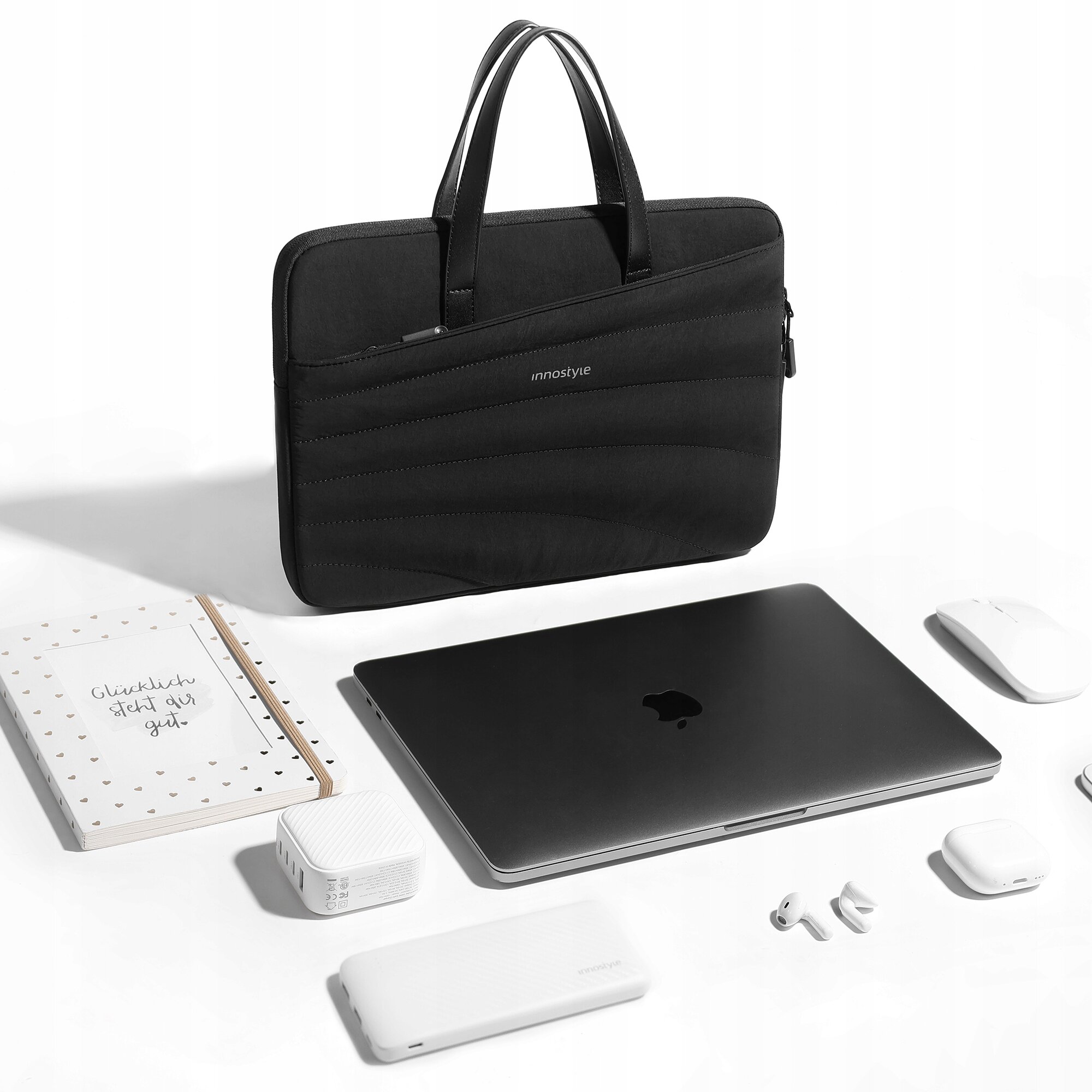 Czarna torba na laptopa z napisem \'innostyle\' znajduje się na białym tle, otoczona laptopem, myszką, notatnikiem z napisem \'Glücklich steht dir gut\', słuchawkami i innymi akcesoriami elektronicznymi.