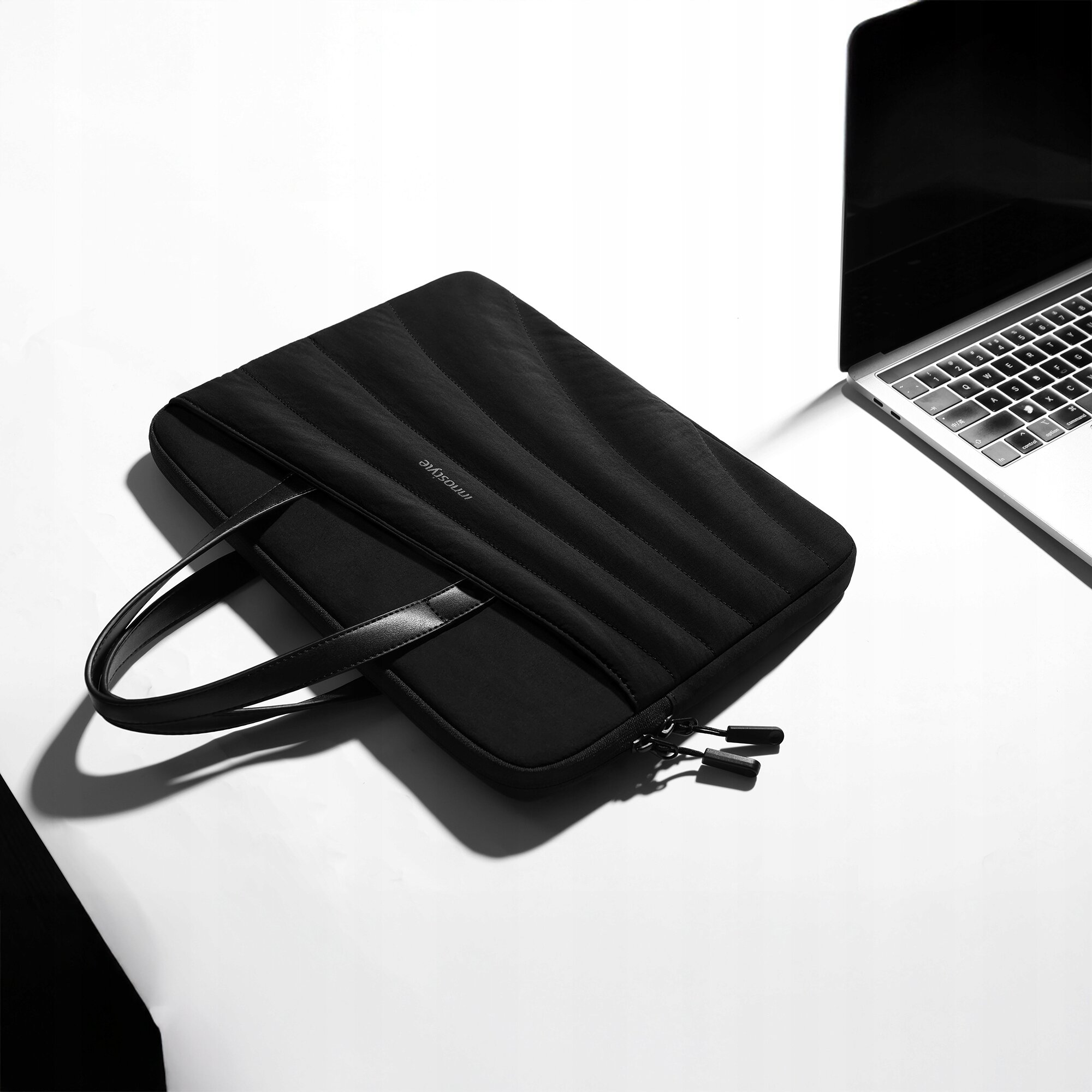 Czarna torba na laptopa z dwoma uchwytami leży na białym tle obok otwartego laptopa. Na torbie widoczny jest napis \'INNOSTYLE\'.
