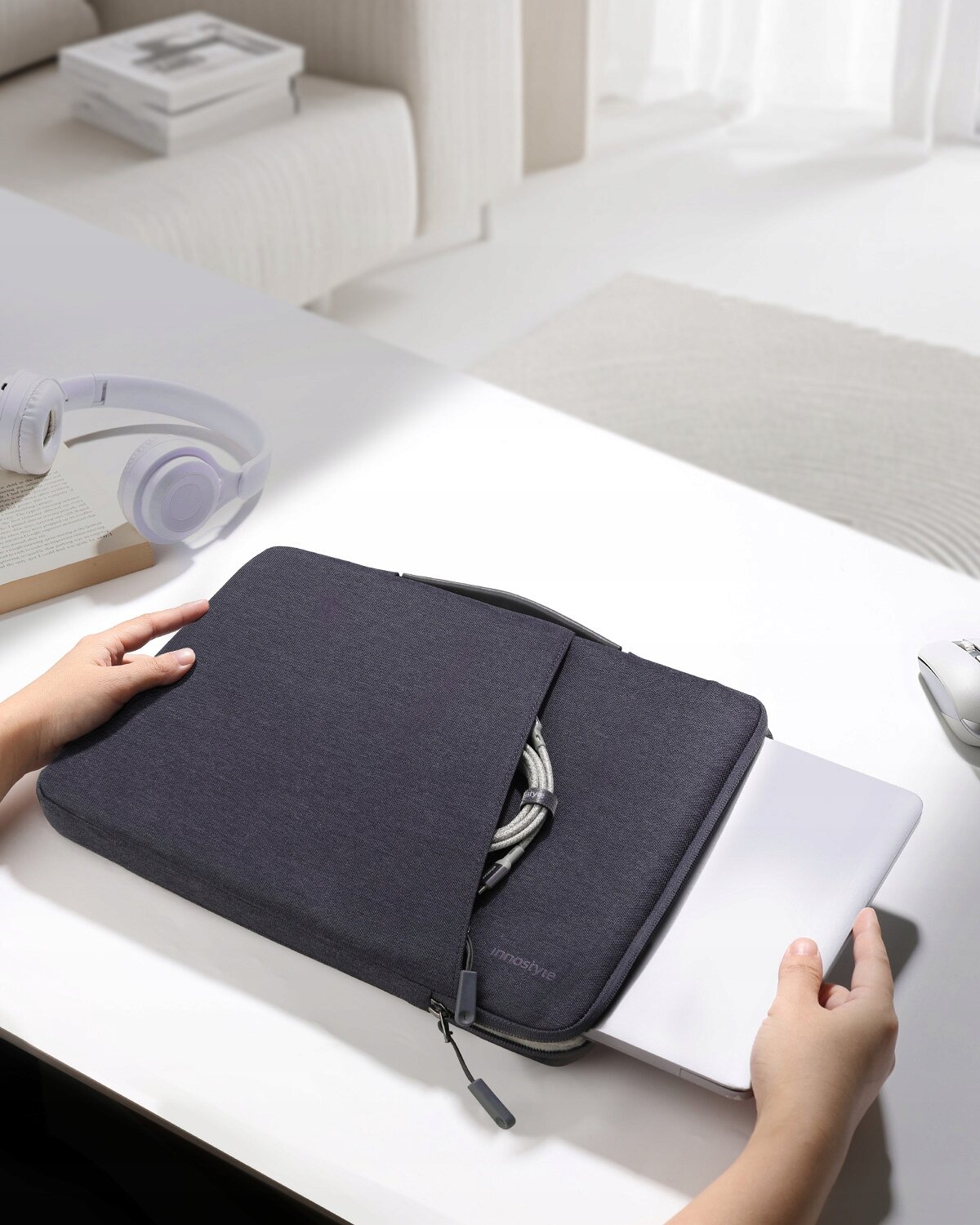 Szara torba na laptopa z napisem \'Innostyle\' na biurku, z laptopem częściowo wsuniętym do środka i widocznymi rękami trzymającymi torbę.