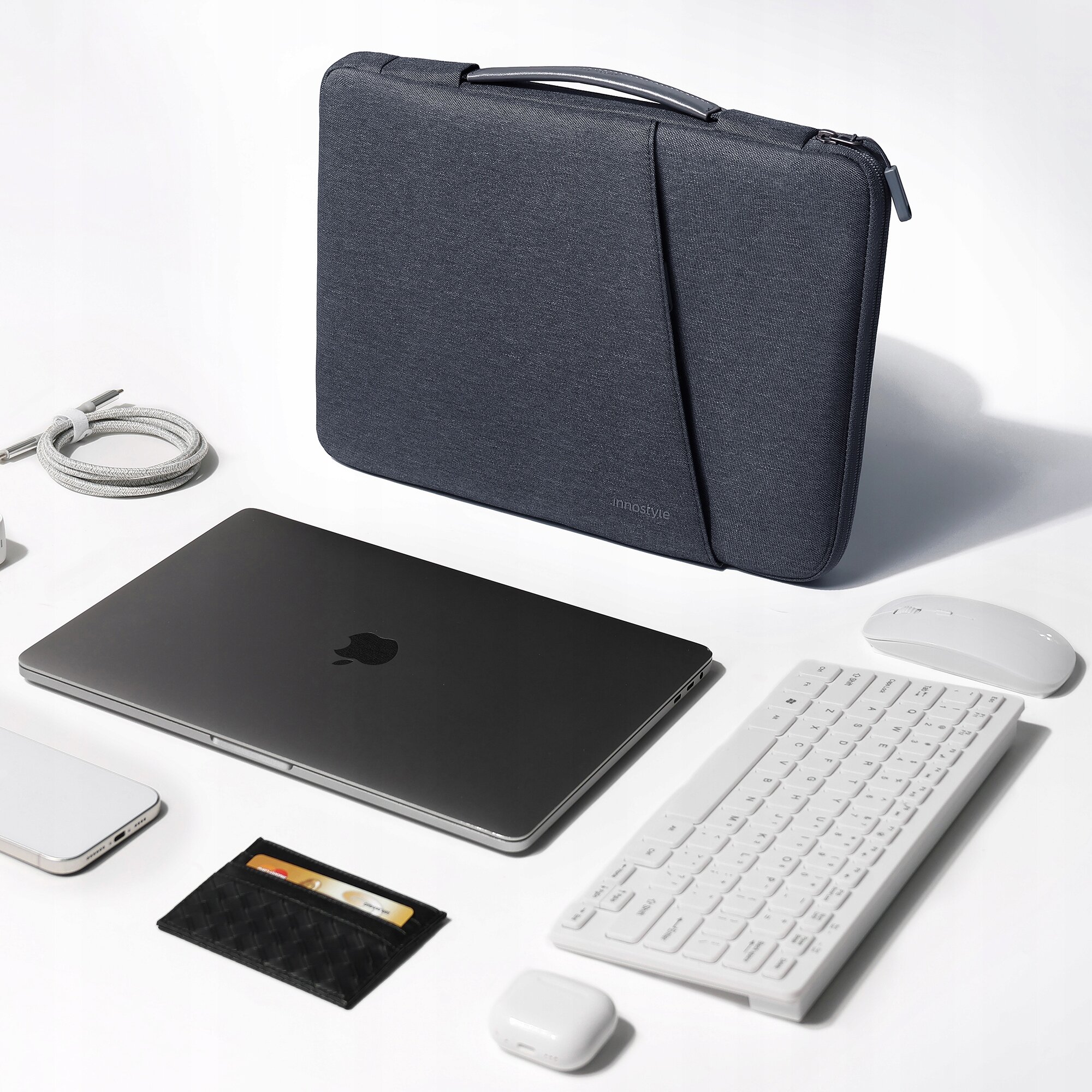 Na obrazie widoczna jest szara torba na laptopa z napisem \'innostyle\', obok której znajdują się laptop, klawiatura, mysz, kabel, telefon, etui na karty i słuchawki. Wszystkie przedmioty są ułożone na białym tle.