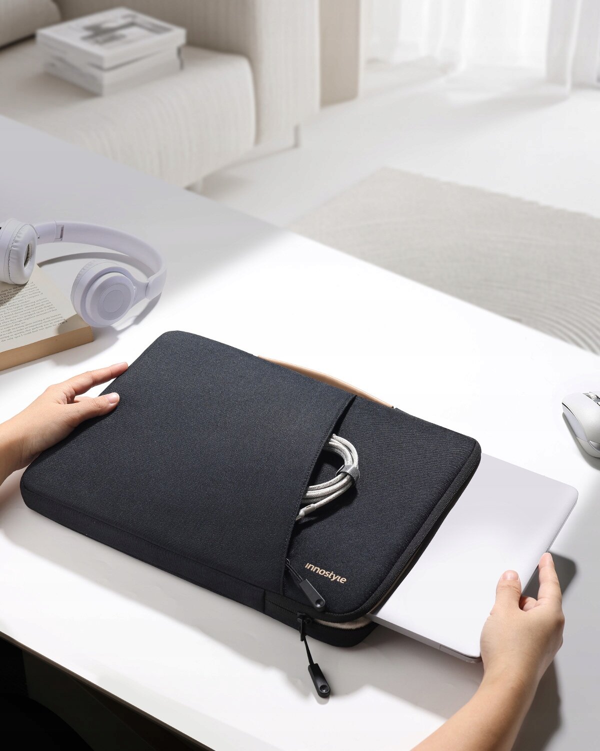Czarna torba na laptopa z napisem \'innostyle\' na biurku, z której wystaje biały laptop i kabel. W tle widoczne są słuchawki, książka i jasne wnętrze pokoju.
