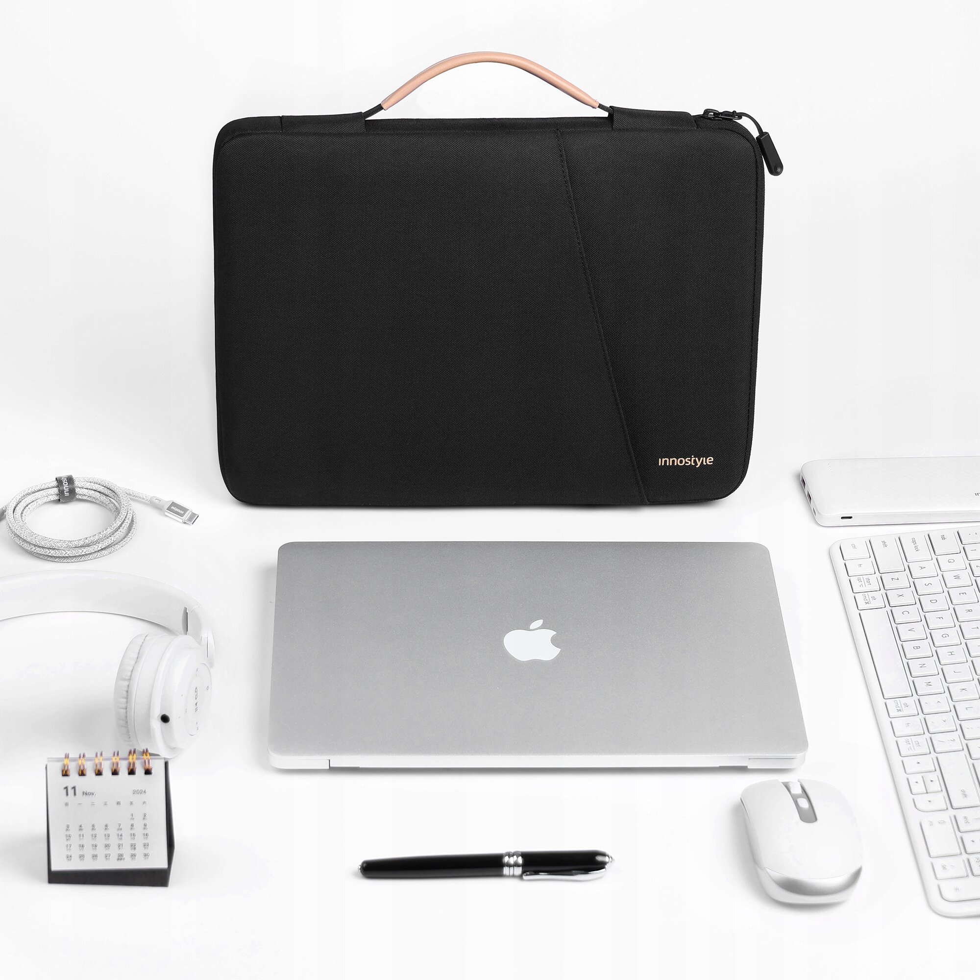 Czarna torba na laptopa z napisem \'innostyle\' leży na białym tle obok laptopa, słuchawek, kalendarza, długopisu, myszki i klawiatury. Torba ma brązowy uchwyt i zamek błyskawiczny.
