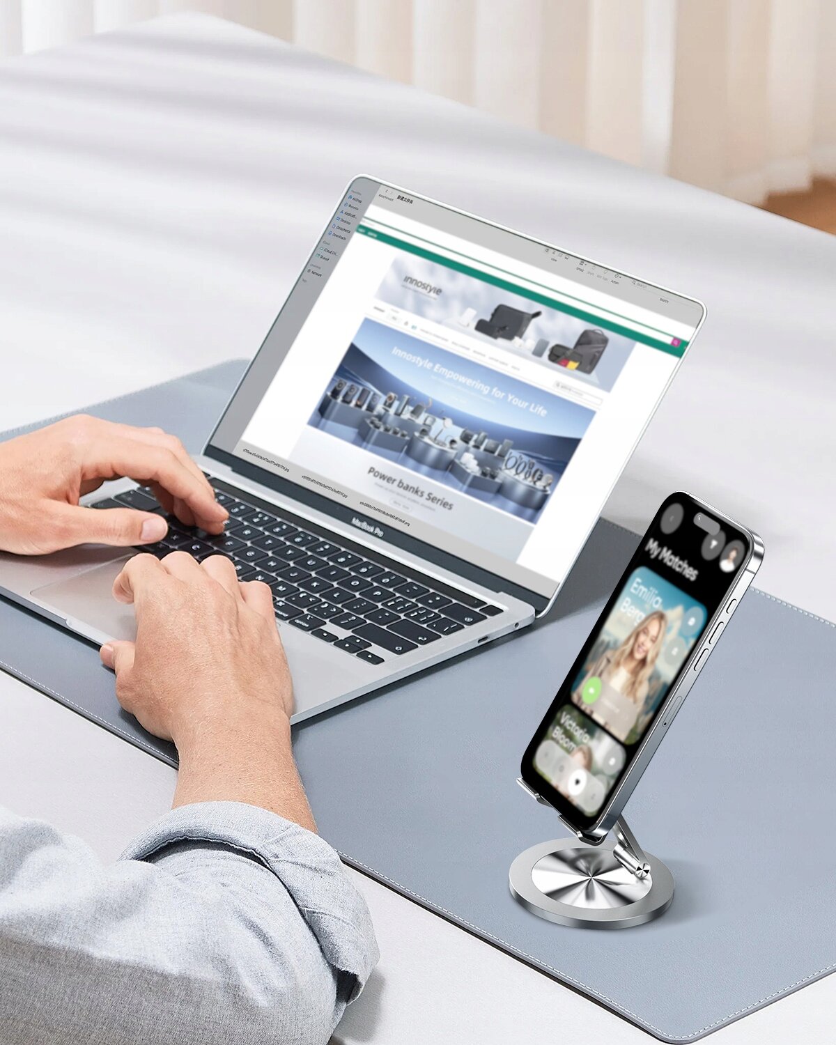 Osoba korzysta z laptopa, a obok na składanym stojaku znajduje się smartfon. Na ekranie laptopa widoczna jest strona internetowa, a na ekranie telefonu aplikacja z muzyką.