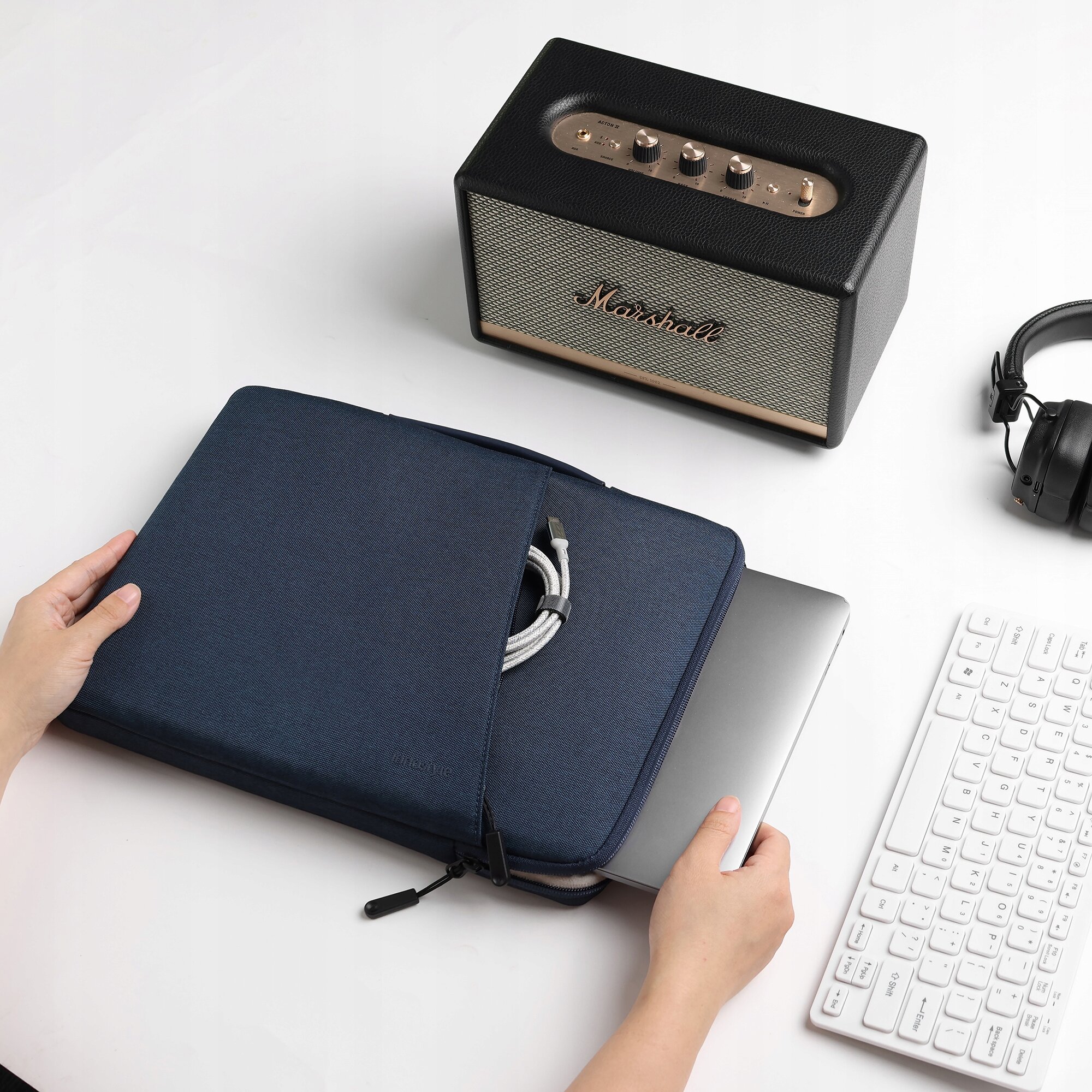 Na zdjęciu widoczna jest niebieska torba na laptopa, z której wystaje laptop oraz kabel, obok znajduje się głośnik z napisem \'Marshall\', klawiatura i słuchawki. Tło jest jednolite i białe.