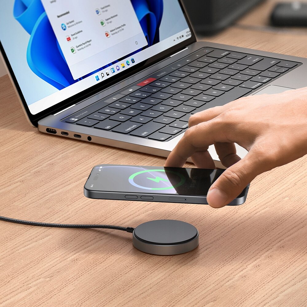 Na drewnianym blacie znajduje się laptop z otwartym ekranem oraz bezprzewodowa ładowarka indukcyjna, na której leży smartfon z widocznym symbolem ładowania. Ręka zbliża się do telefonu, a w tle widać część klawiatury laptopa.