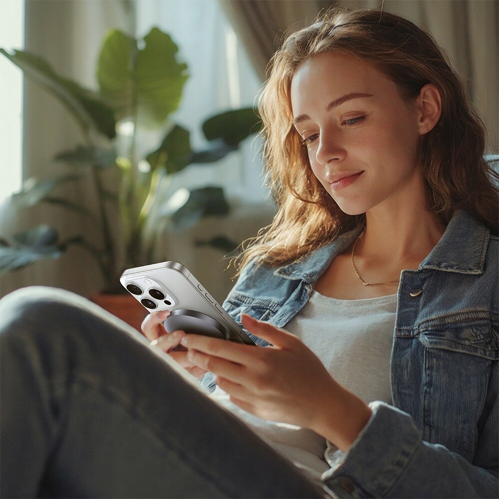 Osoba trzyma smartfon z widoczną ładowarką indukcyjną przymocowaną z tyłu. W tle znajduje się roślina doniczkowa i zasłony.