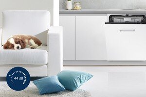Samsung Pewność i komfort. Cichy tryb pracy Wnętrze kuchni z otwartą zmywarką w zabudowie oraz białym fotelem, na którym leży pies. Na podłodze znajdują się dwa niebieskie poduszki, a w lewym dolnym rogu widoczna jest ikona z napisem \'44 dB\'.