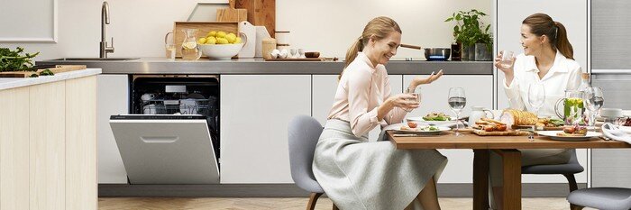 Samsung Jeszcze Wieksza Wygoda Dw60m6050bb 1920 W kuchni przy stole siedzą dwie osoby, rozmawiając i jedząc posiłek. W tle widać otwartą zmywarkę w zabudowie oraz blat z owocami i rośliną.