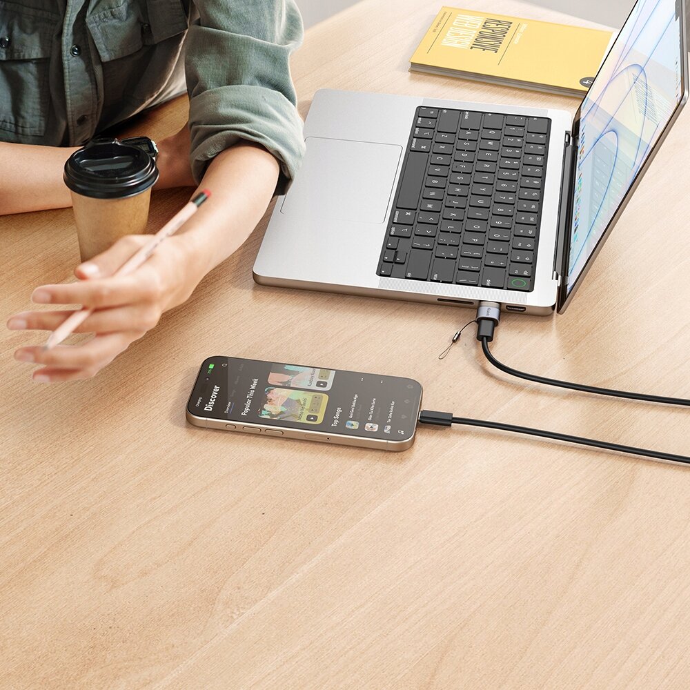 Na drewnianym stole znajduje się otwarty laptop z podłączonym kablem USB-C, obok leży smartfon z włączoną aplikacją muzyczną. Obok laptopa leży żółta książka i kubek z pokrywką, a ręka trzyma ołówek.