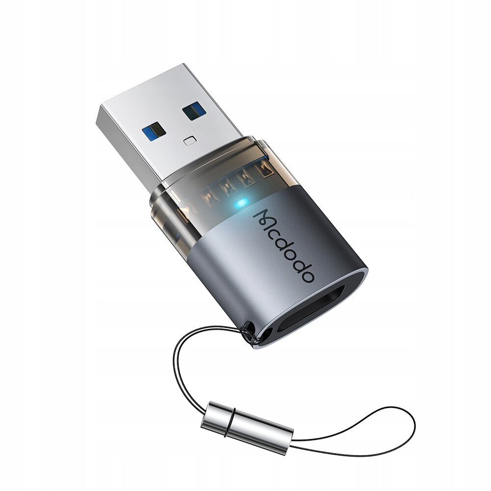 Na obrazie widoczny jest adapter USB z logo \'Mcdodo\', posiadający metaliczną obudowę i przezroczystą część z niebieskim wskaźnikiem LED. Adapter jest wyposażony w smyczkę do łatwego przenoszenia.