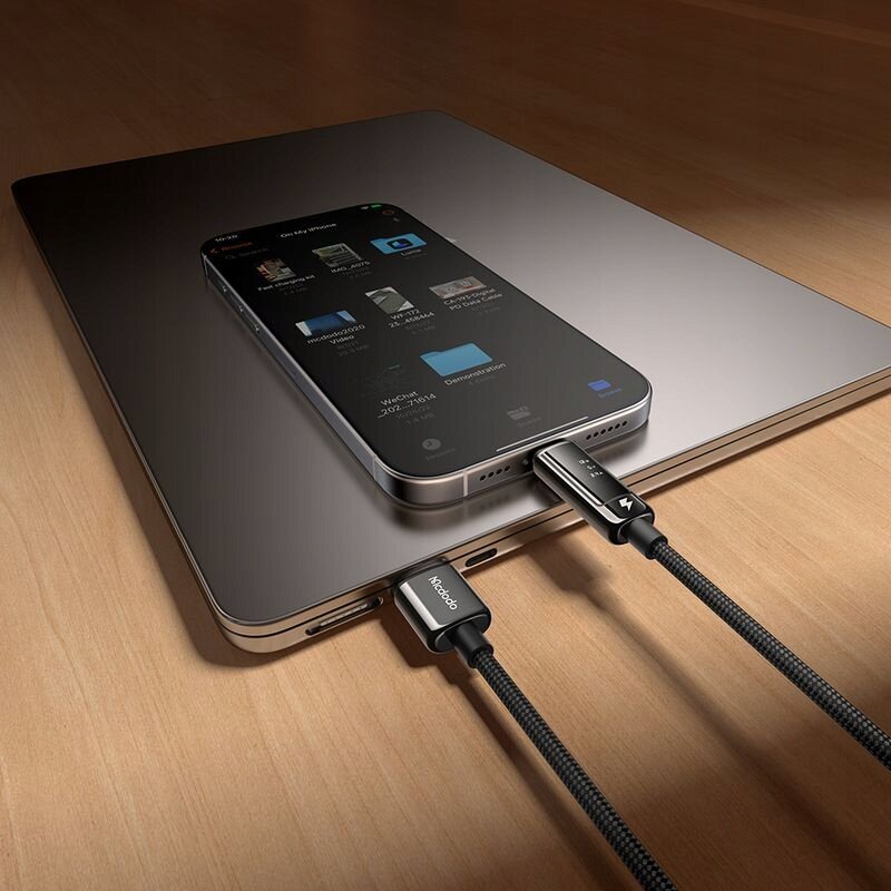 Na drewnianym blacie leży laptop, na którym znajduje się smartfon podłączony do dwóch kabli USB-C. Na kablach widoczne są napisy \'Mcdodo\'.