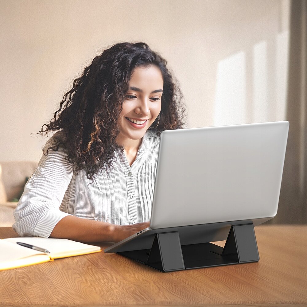 Na zdjęciu znajduje się laptop umieszczony na czarnej, regulowanej podstawce na biurku.