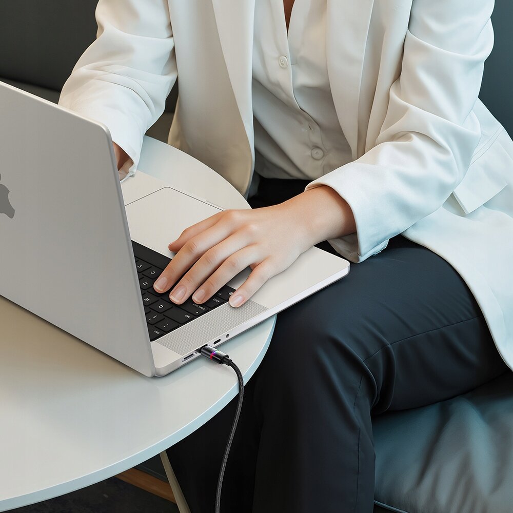 Osoba w białej marynarce i czarnych spodniach używa laptopa na okrągłym stole, do którego podłączony jest czarny kabel USB-C.