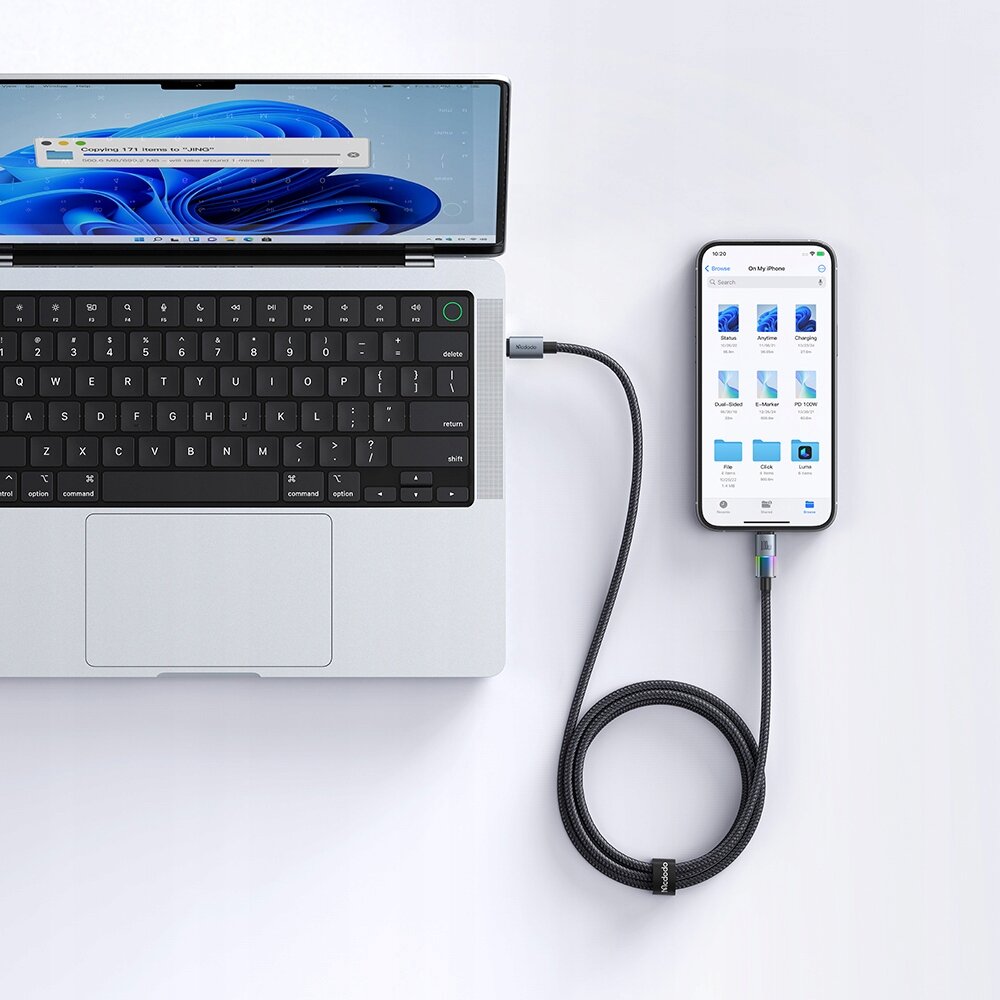 Na białym tle znajduje się laptop z otwartym ekranem i klawiaturą oraz smartfon podłączony do niego czarnym kablem USB-C. Na ekranie smartfona widoczne są ikony aplikacji, a kabel ma plecioną strukturę i świecący wskaźnik LED.