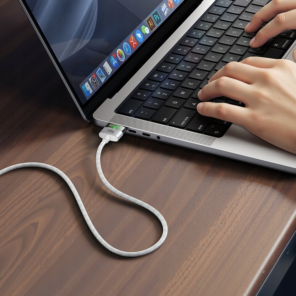 Na zdjęciu widać laptopa na drewnianym stole z podłączonym kablem USB-C Magsafe 3, a na ekranie widoczne są ikony aplikacji. Dłonie osoby piszącej na klawiaturze są również widoczne.