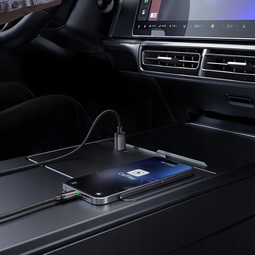W samochodzie na konsoli środkowej leży telefon podłączony do kabla USB, na ekranie widoczny jest napis \'CarPlay\'. W tle widać fragment deski rozdzielczej z ekranem i kratką wentylacyjną.