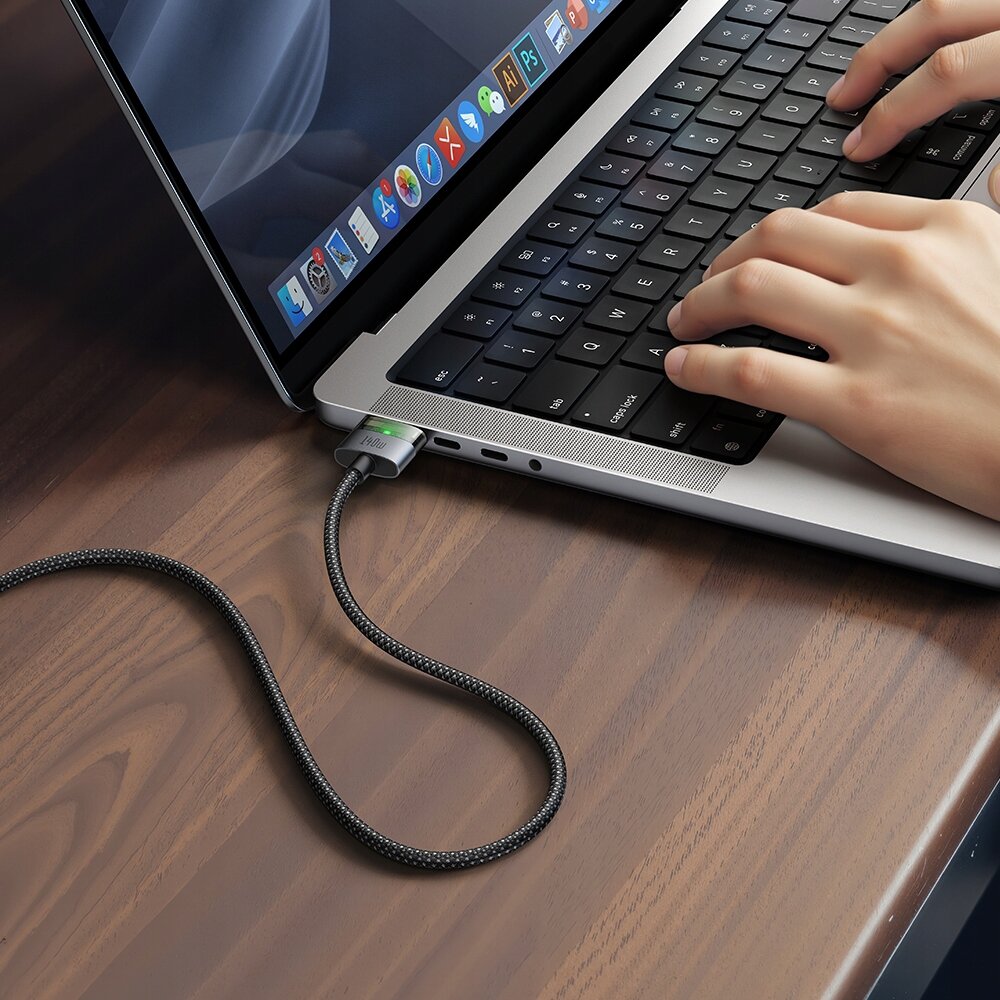 Laptop z podłączonym kablem USB-C Magsafe 3, leżący na drewnianym stole, z widocznymi dłońmi na klawiaturze. Na kablu widoczny jest napis \'Mcdodo\'.