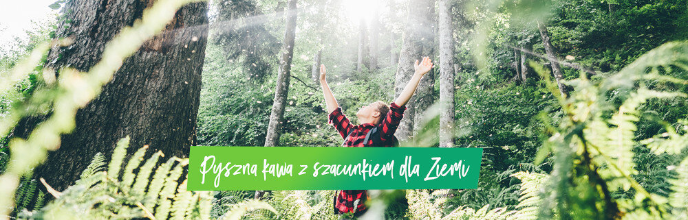 Osoba w czerwono-czarnej koszuli w kratę stoi w lesie z uniesionymi rękami. Na obrazie widnieje tekst: \'Pyszna kawa z szacunkiem dla Ziemi\'.