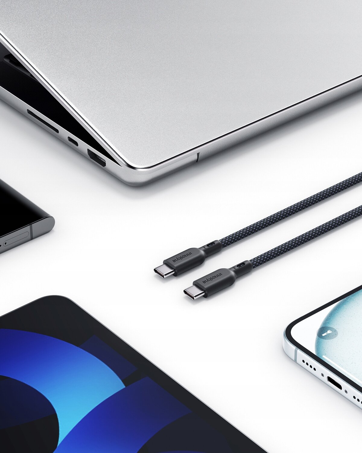 Na zdjęciu widoczny jest kabel USB-C z napisem \'Innostyle\' oraz różne urządzenia elektroniczne na białym tle. Kabel ma plecioną osłonę i jest podłączony do laptopa oraz smartfona.