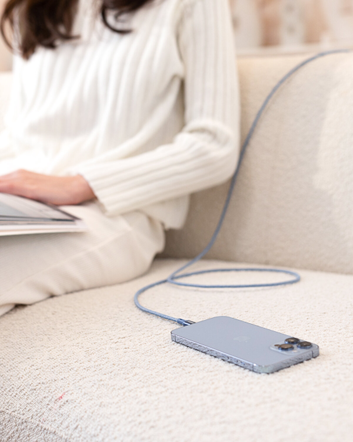 Osoba siedzi na kanapie, trzymając laptopa na kolanach. Obok niej leży telefon podłączony do niebieskiego kabla USB-C Lightning.