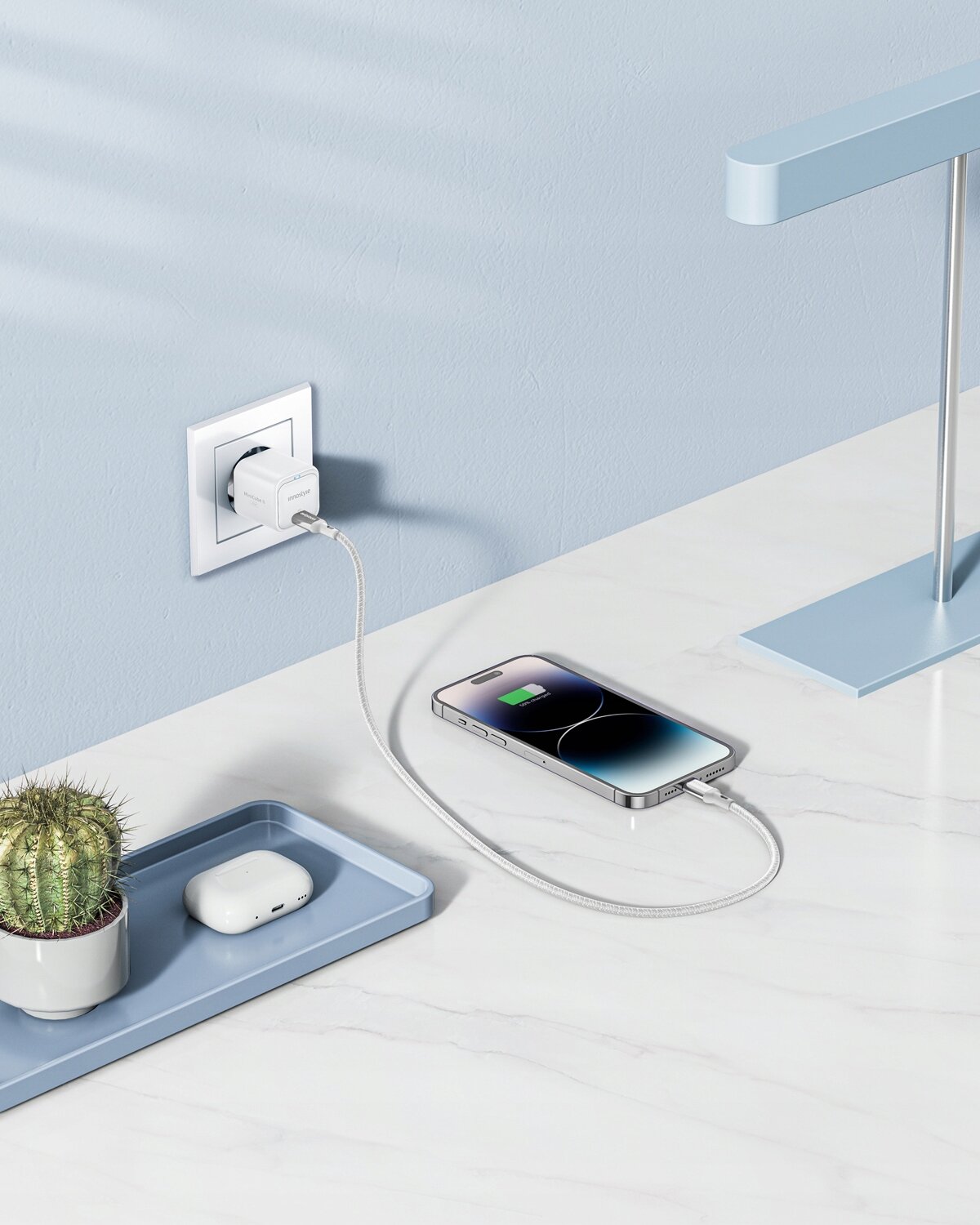 Na białym blacie znajduje się telefon podłączony do ładowarki USB-C, która jest wpięta do gniazdka w ścianie. Obok telefonu leży niebieska taca z kaktusem i słuchawkami bezprzewodowymi.