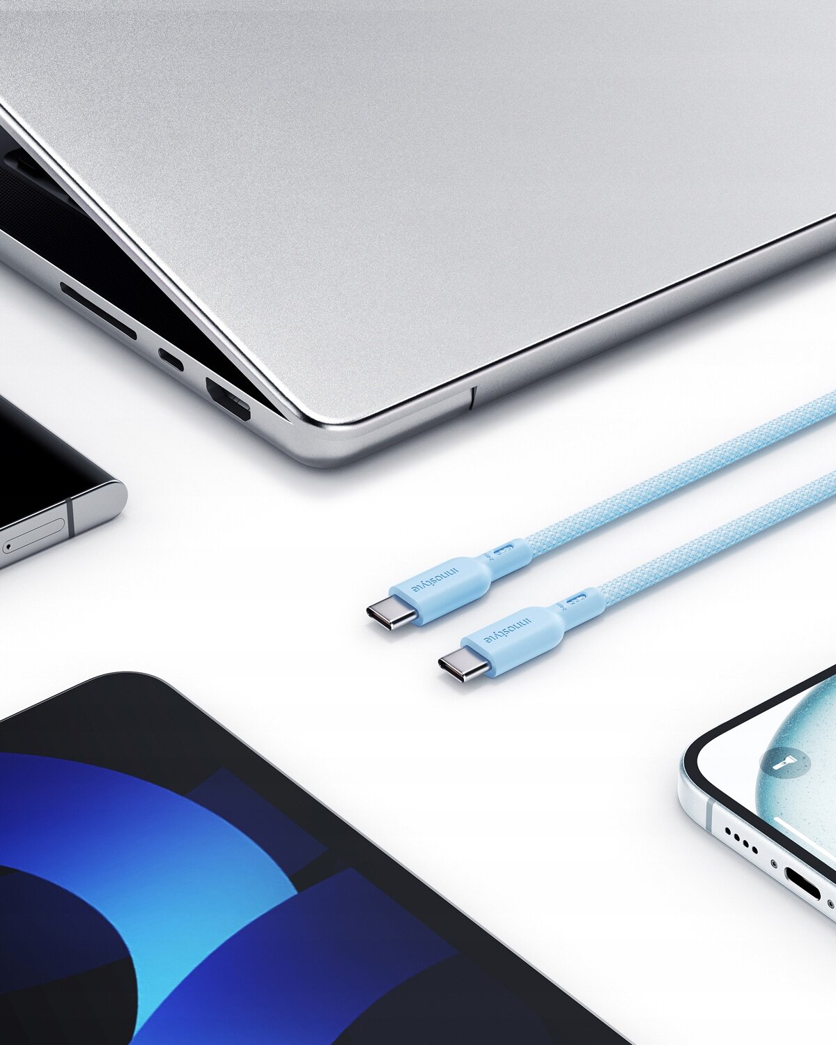Na obrazie widoczne są dwa niebieskie kable USB-C z napisem \'Innostyle\', leżące obok srebrnego laptopa i dwóch smartfonów. Kable mają plecioną strukturę i są przeznaczone do szybkiego ładowania.