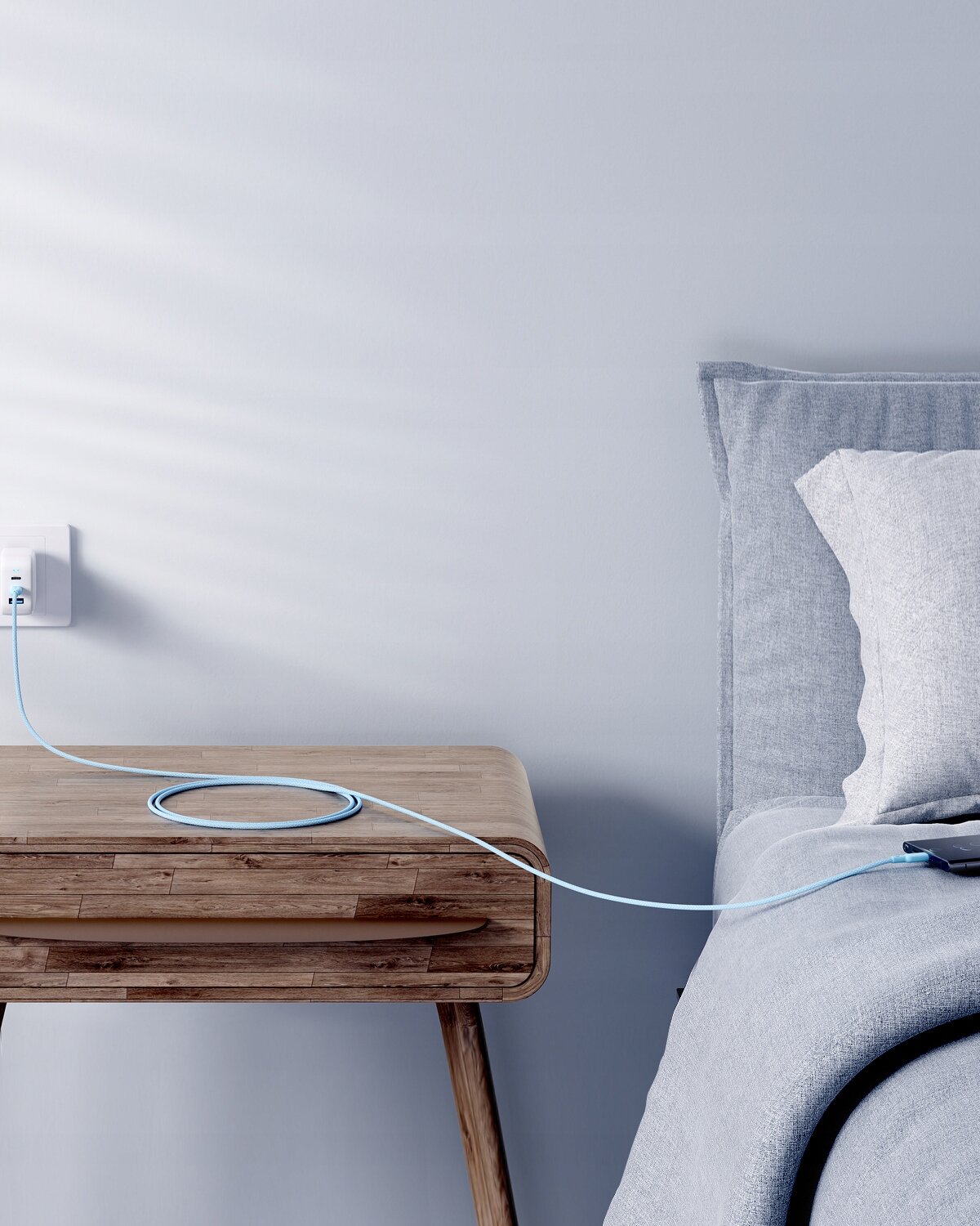 Na zdjęciu widać drewniany stolik nocny z podłączonym do gniazdka kablem USB-C, który biegnie w kierunku łóżka z szarą pościelą. Kabel jest koloru niebieskiego i leży luźno na stoliku.