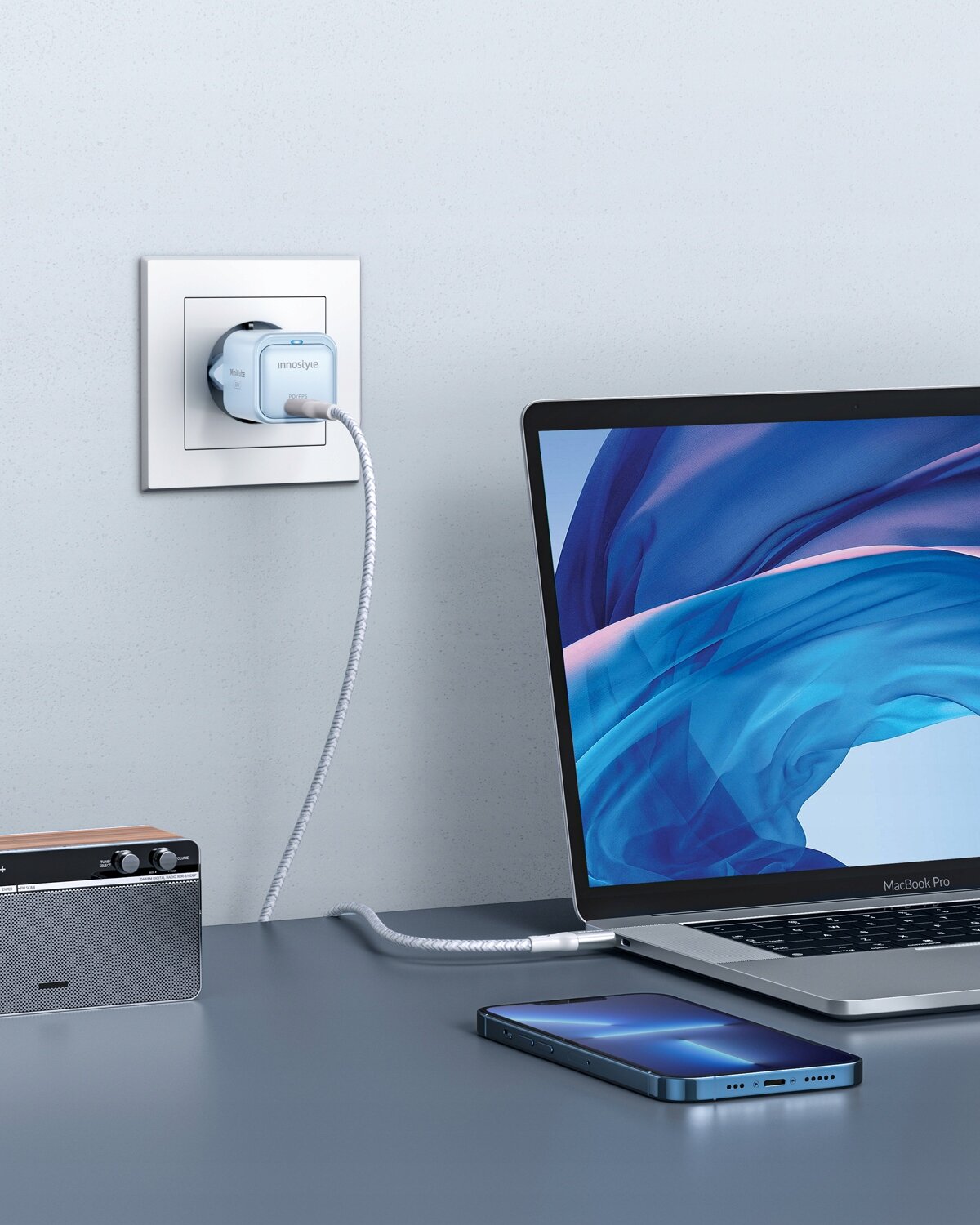 Na obrazie widać ładowarkę USB-C podłączoną do gniazdka ściennego, z napisem \'innostyle\' na obudowie. Obok znajduje się laptop z niebieskim ekranem oraz smartfon leżący na biurku.