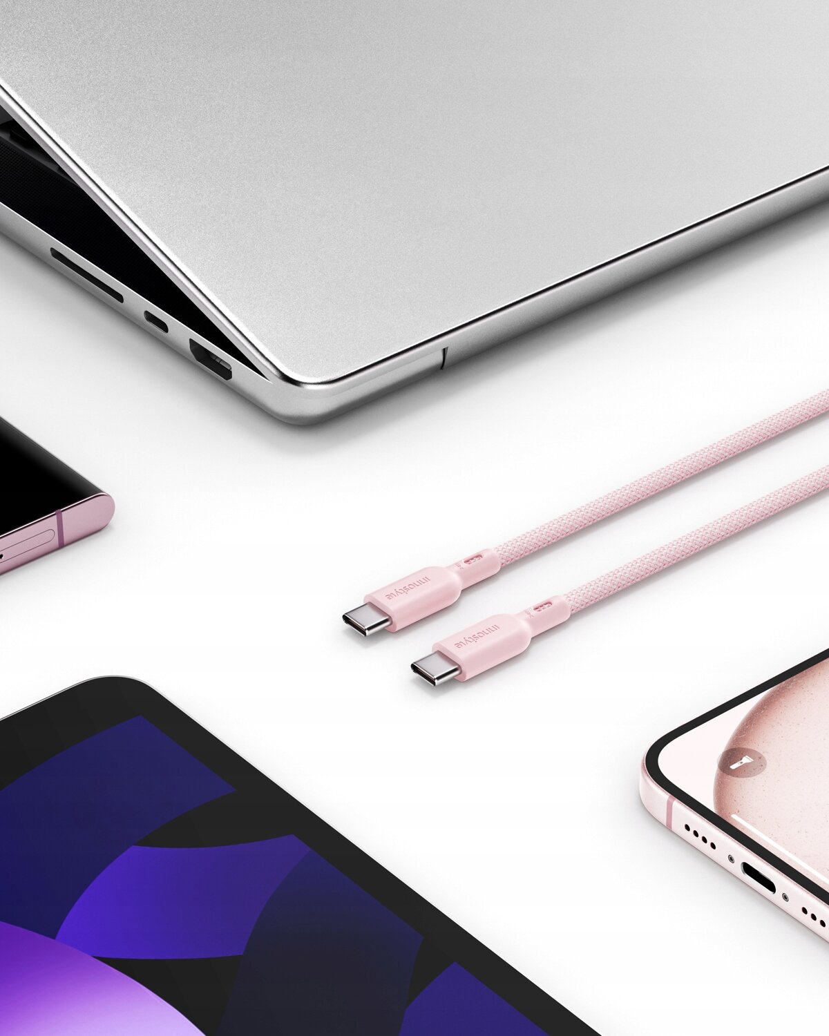 Na obrazie widoczny jest różowy kabel USB-C z napisem \'Innostyle\', otoczony przez różne urządzenia elektroniczne, takie jak laptop i smartfony.
