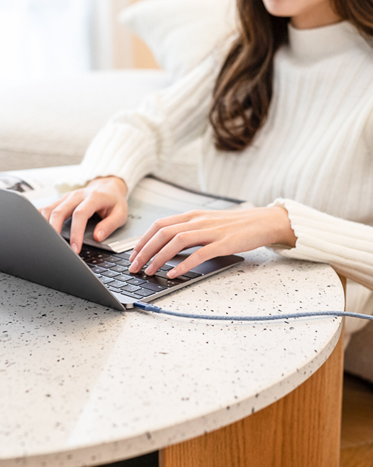 Osoba w białym swetrze pracuje na laptopie przy okrągłym, jasnym stole. Do laptopa podłączony jest niebieski kabel USB-C.