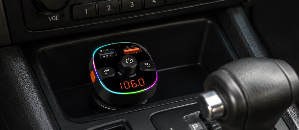 Transmiter FM z wyświetlaczem pokazującym częstotliwość 106.0, otoczony kolorowym podświetleniem, umieszczony w samochodowym uchwycie. Widoczne są przyciski sterujące oraz porty USB oznaczone jako QC 3.0 i 5V/2.4A.