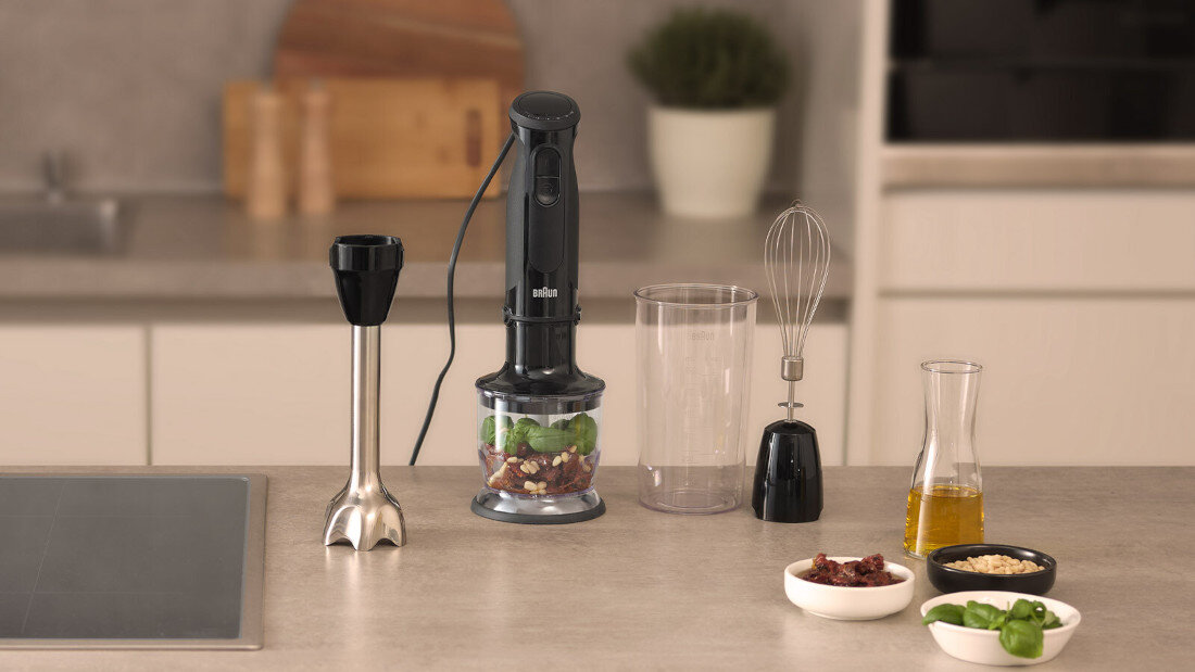 Na blacie kuchennym znajduje się czarny blender ręczny z logo \'braun\', z różnymi akcesoriami, w tym pojemnikiem z pokrojonymi składnikami, trzepaczką i wysokim naczyniem. Obok stoją miseczki z przyprawami i oliwą oraz świeże zioła.