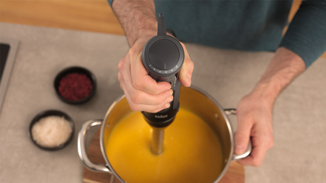 Osoba trzyma czarny blender zanurzeniowy w garnku z żółtą zupą. W tle widoczne są dwie miseczki z przyprawami na blacie.