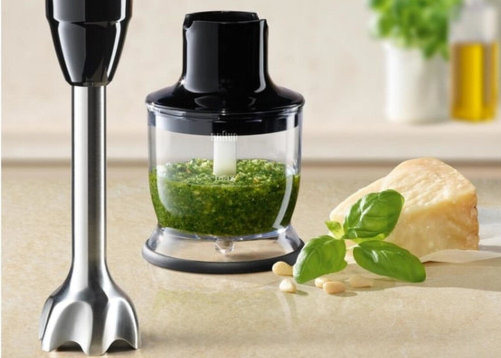 Na blacie kuchennym znajduje się blender ręczny z metalową końcówką oraz pojemnik z czarną pokrywką, wypełniony zieloną masą. Obok leży kawałek sera, liście bazylii i kilka orzeszków.