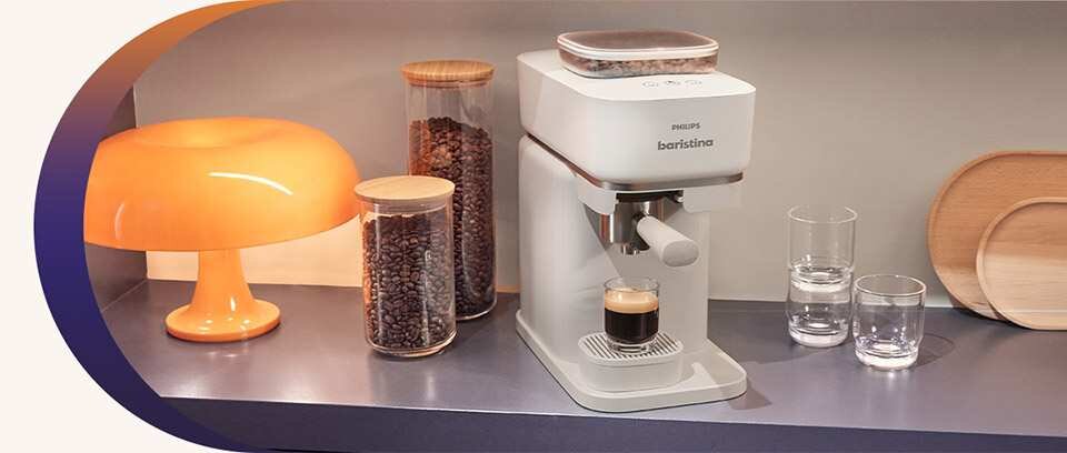 Na blacie kuchennym znajduje się ekspres do kawy PHILIPS Baristina z filiżanką espresso. Obok ekspresu stoją dwa szklane pojemniki z ziarnami kawy, pomarańczowa lampa i dwie szklanki z wodą.